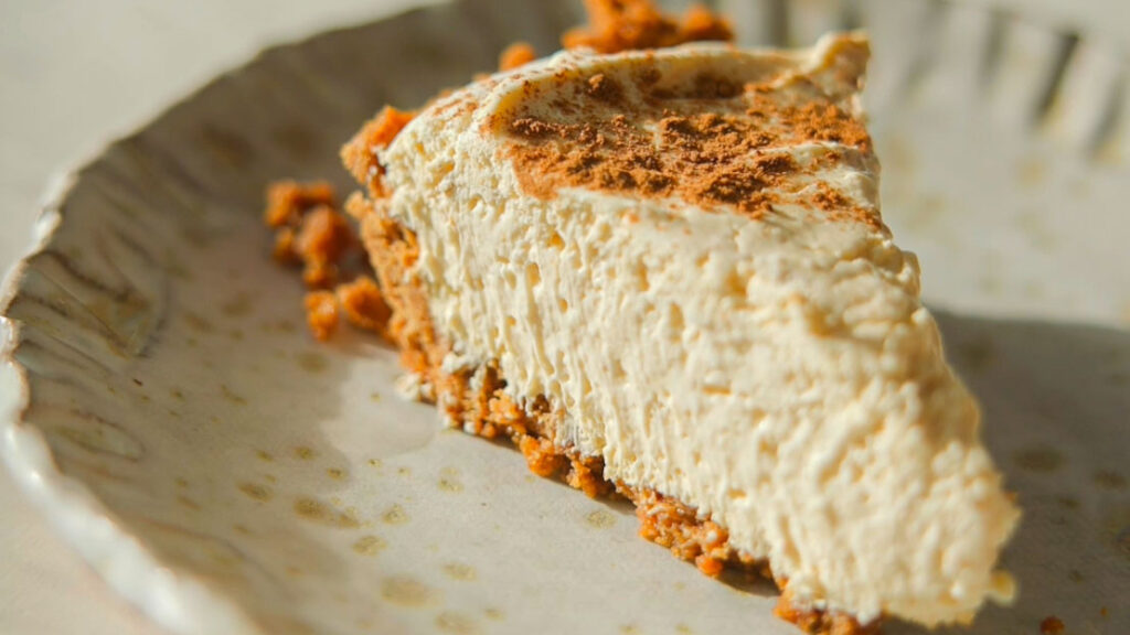 A slice of creamy no-bake pumpkin cheesecake with a graham cracker crust is placed on a decorative ceramic plate. The cheesecake is topped with a dusting of cinnamon, and the image is warmly lit to highlight its texture.